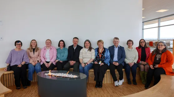 En gruppe på tolv voksne personer sitter på en buet trebenk rundt et rundt ildsted innendørs, i et lyst rom med store vinduer.