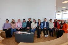 En gruppe på tolv voksne personer sitter på en buet trebenk rundt et rundt ildsted innendørs, i et lyst rom med store vinduer.