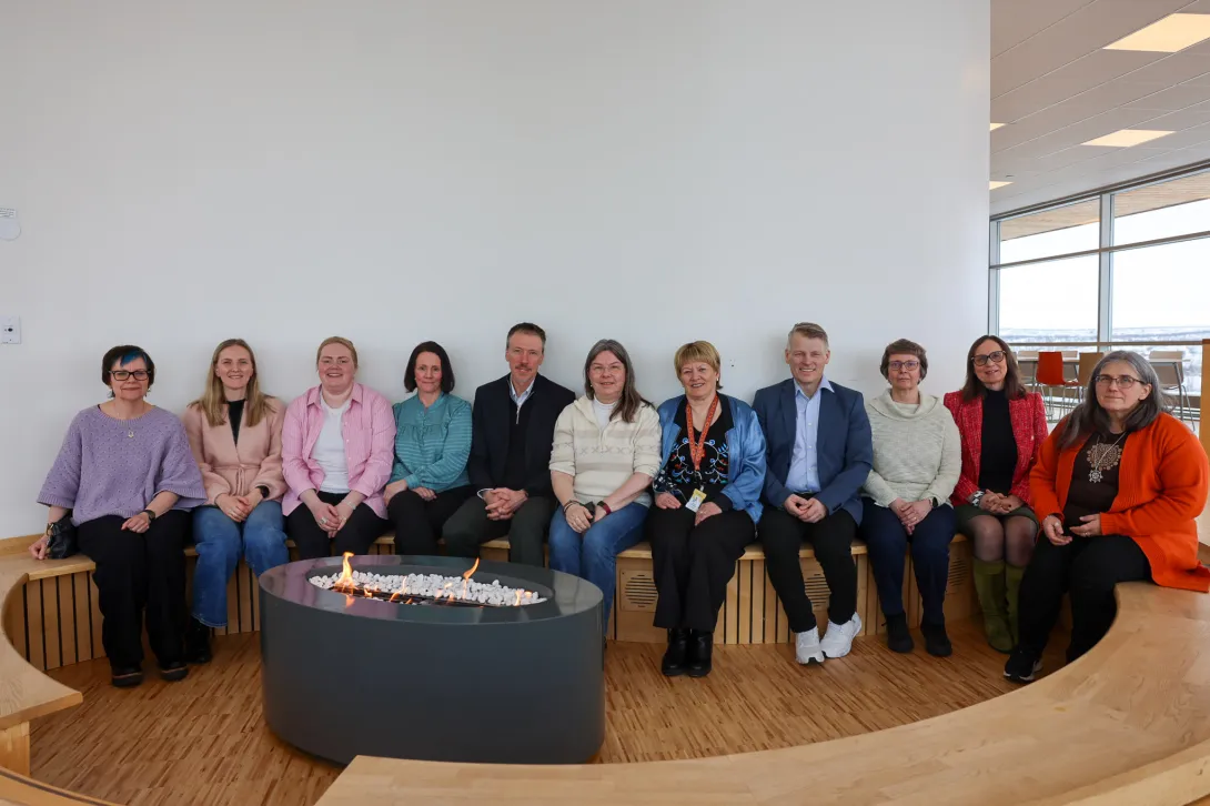 En gruppe på tolv voksne personer sitter på en buet trebenk rundt et rundt ildsted innendørs, i et lyst rom med store vinduer.